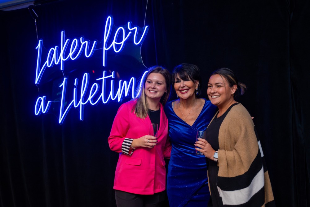 Girls in pink and tan shirts and President Mantella stand in front of Laker for a Lifetime neon sign